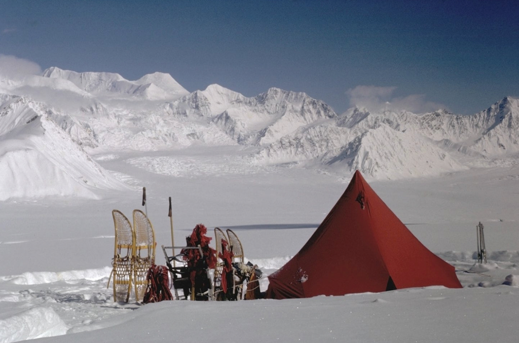 Tent, snowshoes, and supplies on a snow field with snowy mountains behind