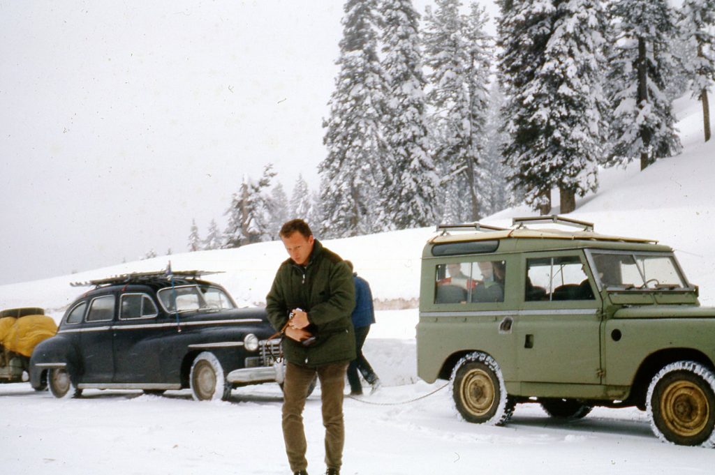Old car with trailer being towed by a Jeep, snow, man walking in front